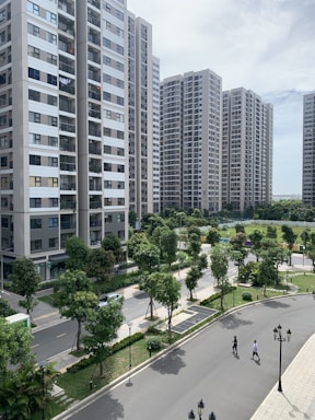 A series of modern high-rise apartment buildings surrounded by well-maintained green spaces and trees. The foreground features a winding road with two people walking, along with classic-style street lamps. The sky is partly cloudy, providing a bright yet calm ambiance.