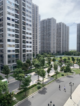 A series of modern high-rise apartment buildings surrounded by well-maintained green spaces and trees. The foreground features a winding road with two people walking, along with classic-style street lamps. The sky is partly cloudy, providing a bright yet calm ambiance.