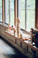 A row of wooden blocks and toys are neatly arranged on a wooden shelf by large windows. The blocks are stacked in various shapes, including a tall tower with a triangular top. The outside scenery through the windows shows greenery blurred in the background.