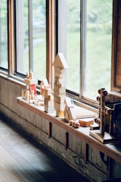 A row of wooden blocks and toys are neatly arranged on a wooden shelf by large windows. The blocks are stacked in various shapes, including a tall tower with a triangular top. The outside scenery through the windows shows greenery blurred in the background.