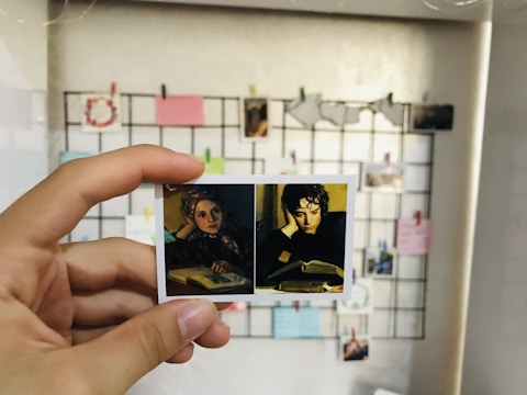 A hand is holding a small printout of two portraits in front of a wall grid filled with various notes and photos. The left portrait features a person wearing a headscarf, looking contemplative, while the right portrait shows a person resting their head on their hand while reading a book.