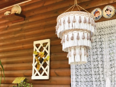 A large decorative hanging made of tiers of long white tassels with beige accents is suspended in a rustic setting. The background features wooden paneling, a lace curtain, a small shelf with a plate, and wall art with floral elements.