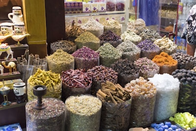 An array of colorful and aromatic spices are displayed in large sacks at what appears to be a spice market. Each sack contains a different type of spice or herb, varying in color and texture. The setting is vibrant with a mix of deep reds, greens, yellows, and browns among other shades. Decorative items and small camel figures are visible on the left side of the image.