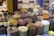 An array of colorful and aromatic spices are displayed in large sacks at what appears to be a spice market. Each sack contains a different type of spice or herb, varying in color and texture. The setting is vibrant with a mix of deep reds, greens, yellows, and browns among other shades. Decorative items and small camel figures are visible on the left side of the image.