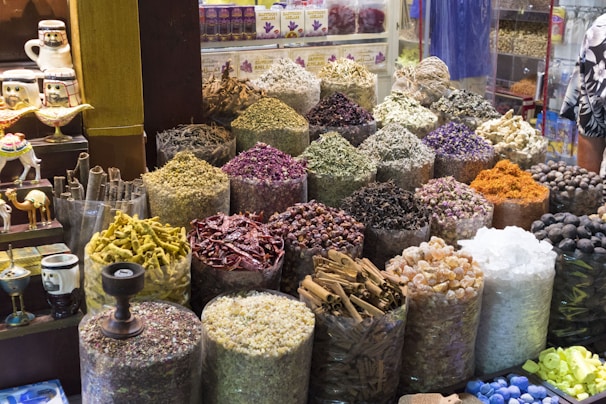 An array of colorful and aromatic spices are displayed in large sacks at what appears to be a spice market. Each sack contains a different type of spice or herb, varying in color and texture. The setting is vibrant with a mix of deep reds, greens, yellows, and browns among other shades. Decorative items and small camel figures are visible on the left side of the image.