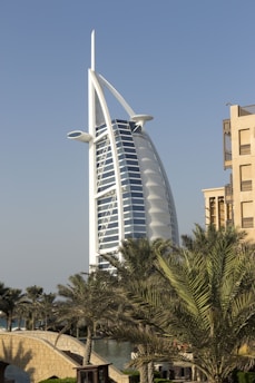 A tall, sail-shaped building with a distinctive modern architecture stands prominently against a clear blue sky. Surrounding the structure are lush palm trees and a small stone bridge, contributing a tropical ambiance to the scene.