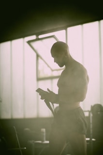 A person with a shaved head stands inside a dimly lit space, likely a gym or training area, wearing dark shorts. The individual appears focused on a smartphone held in their hand, while light streams in from a large window in the background, casting a soft glow.