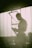 A person with a shaved head stands inside a dimly lit space, likely a gym or training area, wearing dark shorts. The individual appears focused on a smartphone held in their hand, while light streams in from a large window in the background, casting a soft glow.
