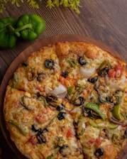 A freshly baked pizza with a variety of toppings, including black olives, sliced green peppers, onions, diced tomatoes, mushrooms, and corn, resting on a wooden board. Nearby, two whole green bell peppers are situated on a wooden table, with some green leaves providing additional decoration.
