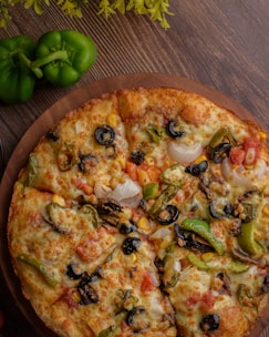 A freshly baked pizza with a variety of toppings, including black olives, sliced green peppers, onions, diced tomatoes, mushrooms, and corn, resting on a wooden board. Nearby, two whole green bell peppers are situated on a wooden table, with some green leaves providing additional decoration.