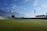 A large open cricket stadium with green grass covering the field. Mountains are visible in the background under a blue sky with scattered clouds. The stadium is empty, with colorful seating in the stands and tall floodlights towering over the field.