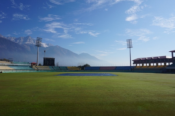 A large open cricket stadium with green grass covering the field. Mountains are visible in the background under a blue sky with scattered clouds. The stadium is empty, with colorful seating in the stands and tall floodlights towering over the field.