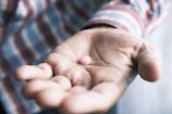 A hand is open with the palm facing upwards, holding a single pink pill. The person is wearing a plaid, long-sleeved shirt. The focus is on the pill, with the background softly blurred.