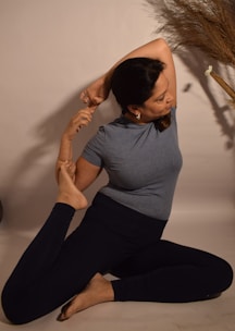 A person practices a seated yoga pose, stretching one arm over their head and holding one foot with the other hand. The background is neutral, with a decorative dried plant on one side.