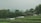 Two small wooden houses with green roofs are nestled amidst lush greenery and terraced fields. White tents or canopies are scattered in front of the structures. Dense trees surround the area, and a hazy mountain can be seen in the background.