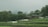 Two small wooden houses with green roofs are nestled amidst lush greenery and terraced fields. White tents or canopies are scattered in front of the structures. Dense trees surround the area, and a hazy mountain can be seen in the background.
