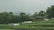 Two small wooden houses with green roofs are nestled amidst lush greenery and terraced fields. White tents or canopies are scattered in front of the structures. Dense trees surround the area, and a hazy mountain can be seen in the background.