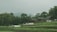 Two small wooden houses with green roofs are nestled amidst lush greenery and terraced fields. White tents or canopies are scattered in front of the structures. Dense trees surround the area, and a hazy mountain can be seen in the background.