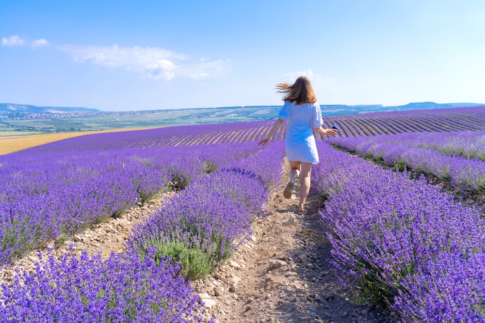 woman in white dress walking on dirt pathway between purple flower fields during daytime