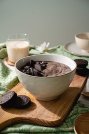 A bowl filled with cookies and cream flavored dessert topped with crushed cookies is placed on a wooden board. Surrounding it are whole cookies, a glass of milk, and a cup on a saucer. The setup rests on a green and white patterned tablecloth.