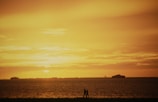 A couple walks hand in hand along the beach against the backdrop of a stunning sunset over the ocean, with large ships visible on the horizon. The sky is painted in warm hues of orange and yellow, reflecting on the tranquil water.