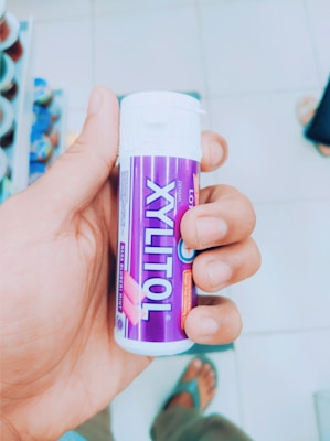 A hand holds a small cylindrical container with a purple label that reads 'Xylitol' and 'Rasa Bubblemint'. The background shows rows of shelves, indicating a store setting.