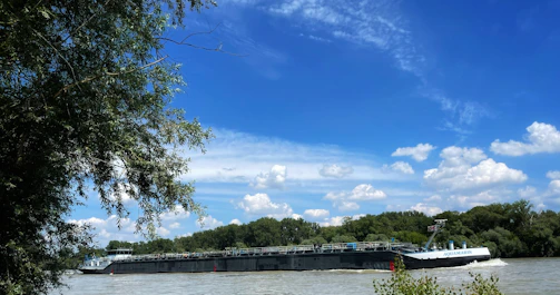 A barge travels on a wide river surrounded by lush green trees under a bright blue sky with scattered white clouds. The calm water contrasts with the vibrant greenery and the solid structure of the barge.