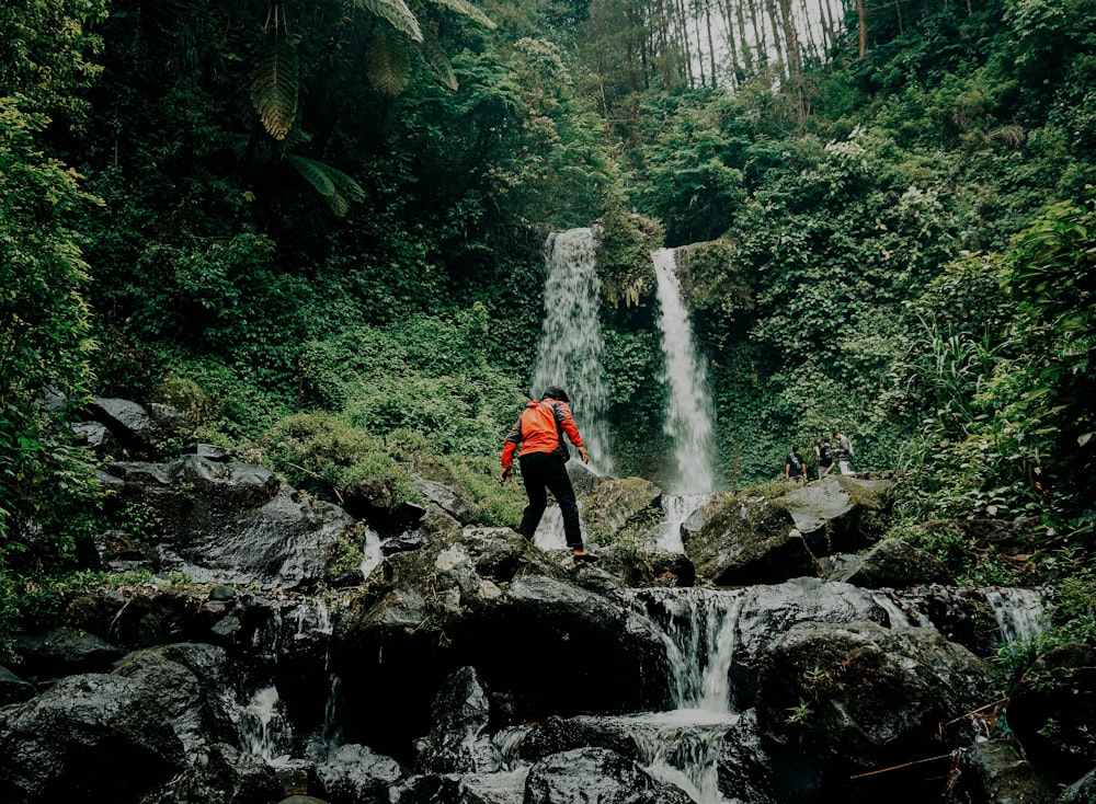 man in red jacket standing on rocky river during daytime