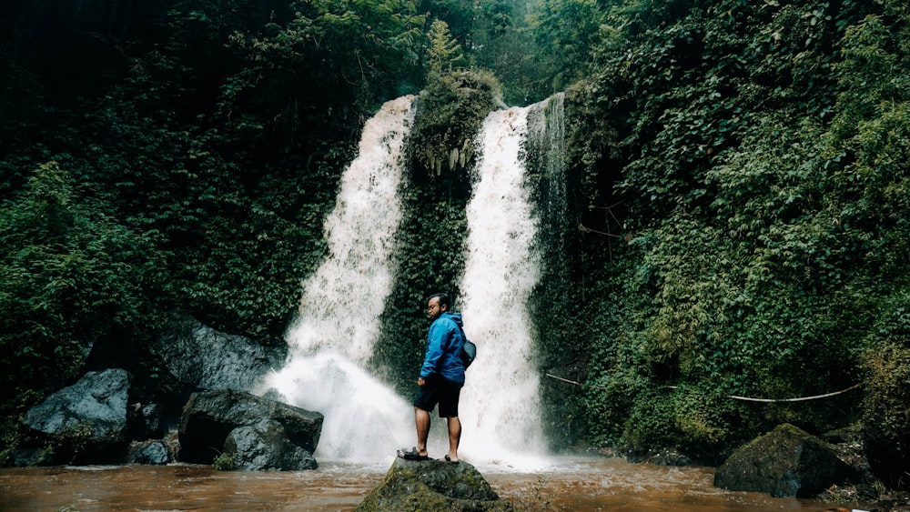 man in blue jacket standing on rock near waterfalls during daytime