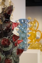 Intricately designed glass sculptures with vibrant colors are displayed on pedestals. The foreground features a dark sculpture with red and translucent spikes. In the background, blue and yellow glass sculptures add contrast and depth.