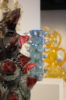 Intricately designed glass sculptures with vibrant colors are displayed on pedestals. The foreground features a dark sculpture with red and translucent spikes. In the background, blue and yellow glass sculptures add contrast and depth.