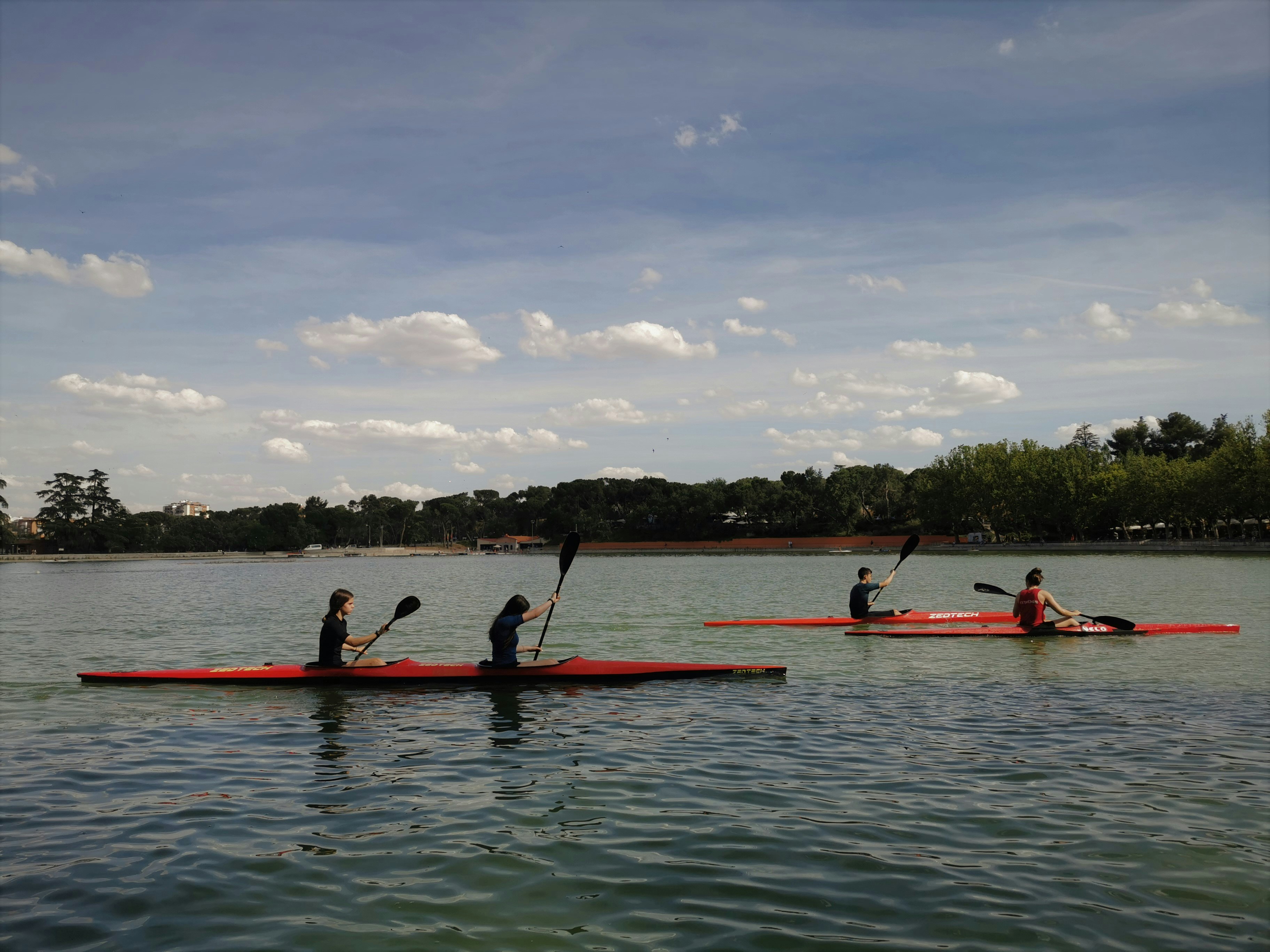 Four people paddle two red kayaks across a calm lake.
