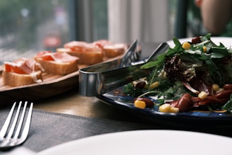 A fresh salad with greens, corn, and kidney beans is presented on a dark blue patterned plate, accompanied by a pair of metal tongs. In the background, there are slices of bread topped with tomato and a slice of ham, arranged neatly on a wooden board. The setting appears to be a dining table with a fork positioned at the foreground.