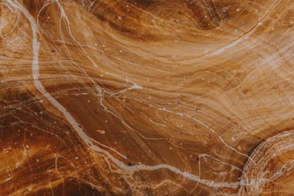 A close-up view of a polished surface with swirling patterns and textures in shades of brown and beige. Thin white lines meander across the surface, resembling natural stone or wood grain.