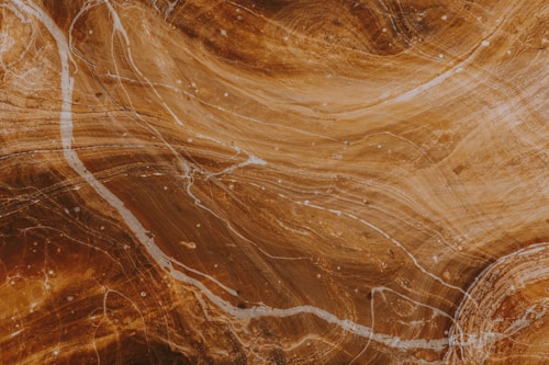 A close-up view of a polished surface with swirling patterns and textures in shades of brown and beige. Thin white lines meander across the surface, resembling natural stone or wood grain.