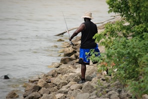 A person wearing a black sleeveless shirt, blue shorts, and a beige hat is fishing while standing on a rocky shoreline. The person holds a fishing rod, and nearby, there is foliage including green bushes.