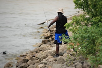 A person wearing a black sleeveless shirt, blue shorts, and a beige hat is fishing while standing on a rocky shoreline. The person holds a fishing rod, and nearby, there is foliage including green bushes.