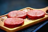 Three raw beef burger patties are placed on a wooden cutting board. The patties appear seasoned with salt and pepper, and the board is set against a dark background. Coarse salt and peppercorns are scattered around the board.