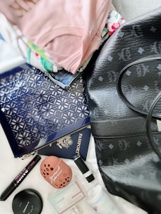 A collection of personal items spread out on a surface including a black leather bag with logos, a blue patterned clutch, a passport, a small round makeup compact, a pen, a lipstick, folded clothing with floral patterns, and various small accessories.