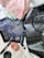 A collection of personal items spread out on a surface including a black leather bag with logos, a blue patterned clutch, a passport, a small round makeup compact, a pen, a lipstick, folded clothing with floral patterns, and various small accessories.
