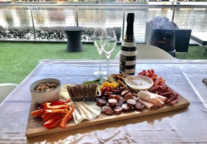 A charcuterie board arranged with a variety of meats, cheeses, crackers, vegetables, and dips on a wooden platter placed on a table. A bottle of wine and two empty wine glasses are positioned behind the board. The setting overlooks a body of water with outdoor seating and greenery, creating a casual and inviting ambiance.