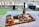 A charcuterie board arranged with a variety of meats, cheeses, crackers, vegetables, and dips on a wooden platter placed on a table. A bottle of wine and two empty wine glasses are positioned behind the board. The setting overlooks a body of water with outdoor seating and greenery, creating a casual and inviting ambiance.