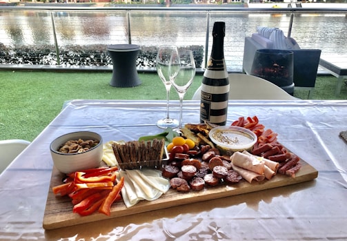 A charcuterie board arranged with a variety of meats, cheeses, crackers, vegetables, and dips on a wooden platter placed on a table. A bottle of wine and two empty wine glasses are positioned behind the board. The setting overlooks a body of water with outdoor seating and greenery, creating a casual and inviting ambiance.
