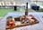 A charcuterie board arranged with a variety of meats, cheeses, crackers, vegetables, and dips on a wooden platter placed on a table. A bottle of wine and two empty wine glasses are positioned behind the board. The setting overlooks a body of water with outdoor seating and greenery, creating a casual and inviting ambiance.