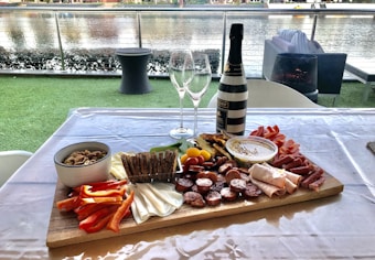 A charcuterie board arranged with a variety of meats, cheeses, crackers, vegetables, and dips on a wooden platter placed on a table. A bottle of wine and two empty wine glasses are positioned behind the board. The setting overlooks a body of water with outdoor seating and greenery, creating a casual and inviting ambiance.