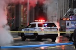 A police vehicle with flashing lights is parked near an illuminated emergency services truck, surrounded by caution tape on a wet urban street. Thick steam rises on the left side of the image, obscuring part of the scene.