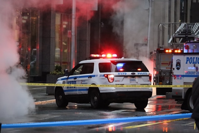A police vehicle with flashing lights is parked near an illuminated emergency services truck, surrounded by caution tape on a wet urban street. Thick steam rises on the left side of the image, obscuring part of the scene.