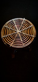 A circular woven object with concentric rings made of rattan or bamboo material is placed against a dark background. It has a sturdy, handcrafted appearance with a detailed weaving pattern evident in the intersecting lines.