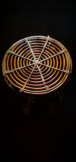 A circular woven object with concentric rings made of rattan or bamboo material is placed against a dark background. It has a sturdy, handcrafted appearance with a detailed weaving pattern evident in the intersecting lines.
