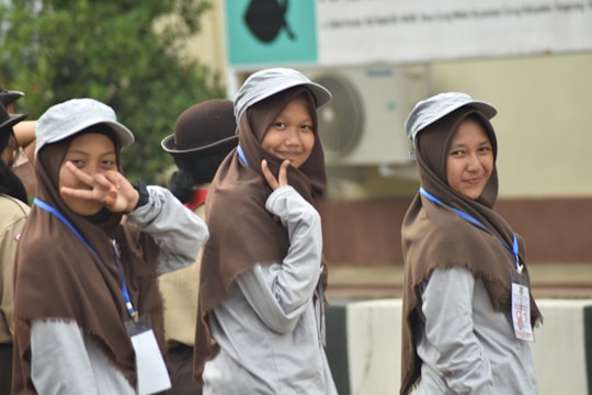 Three young women wearing uniforms with brown hijabs and gray caps are smiling and posing for the camera. Two of them make peace signs, and they have lanyards with ID cards around their necks. The setting appears to be an outdoor event, possibly linked to scouts or a similar organization. Behind them is a blurred background with greenery and a building with an air conditioning unit.